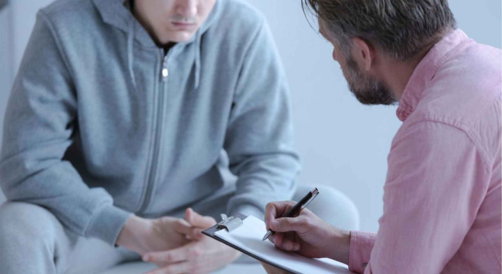 Counselor taking notes on a clipboard while listening to a young man during a therapy session