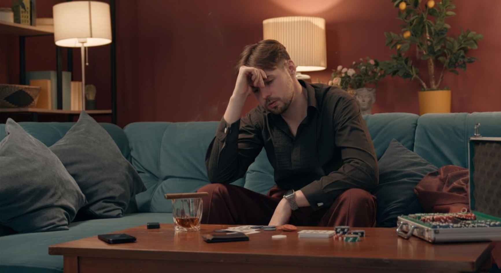 Distressed man sitting on a couch with poker chips and a drink on the table in front of him