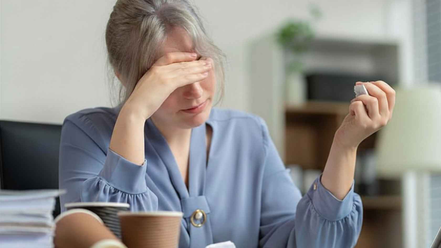 Exhausted woman rubbing her eyes at a desk surrounded by paperwork and coffee cups