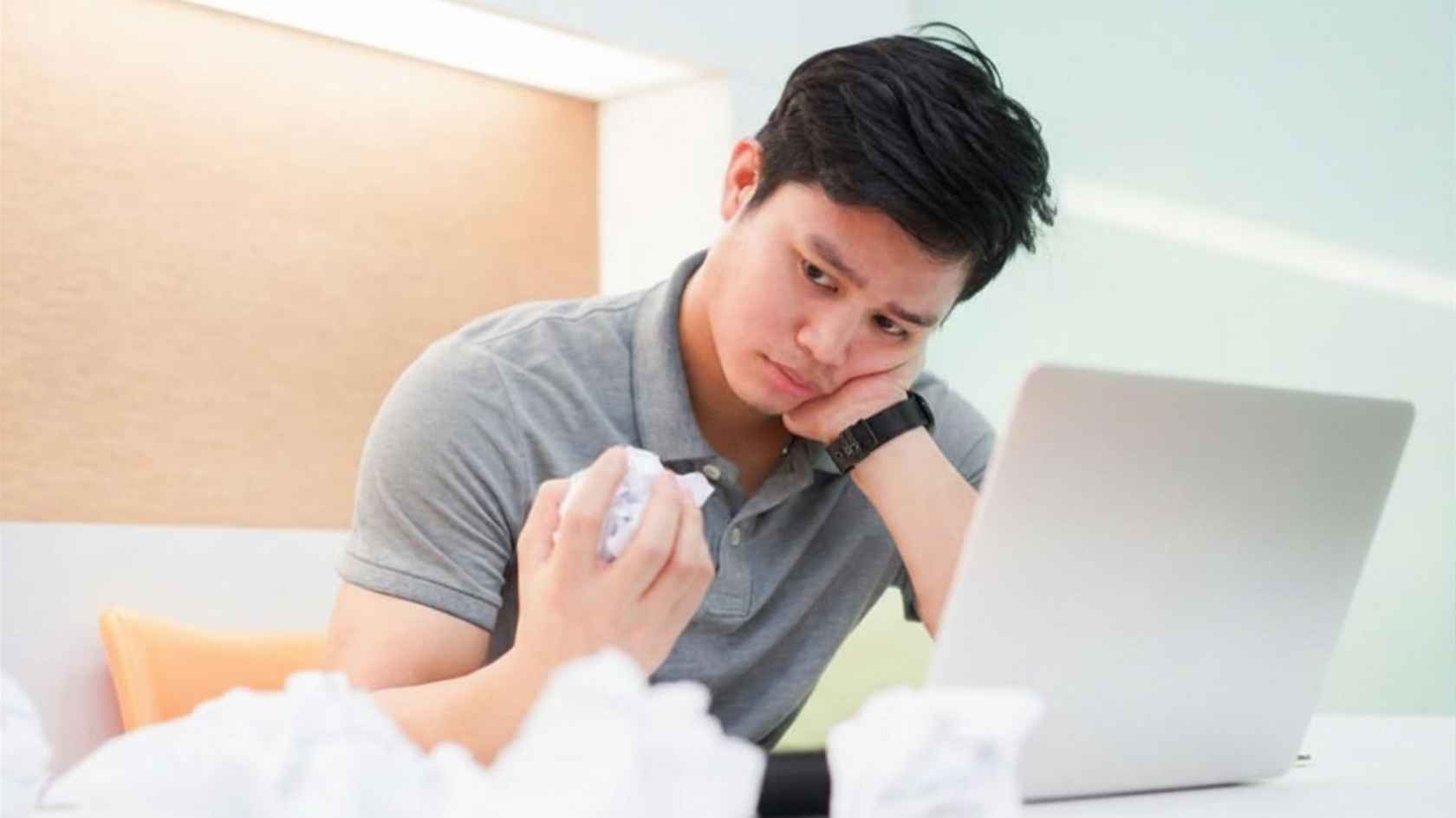 Frustrated young man at a laptop surrounded by crumpled paper on his desk