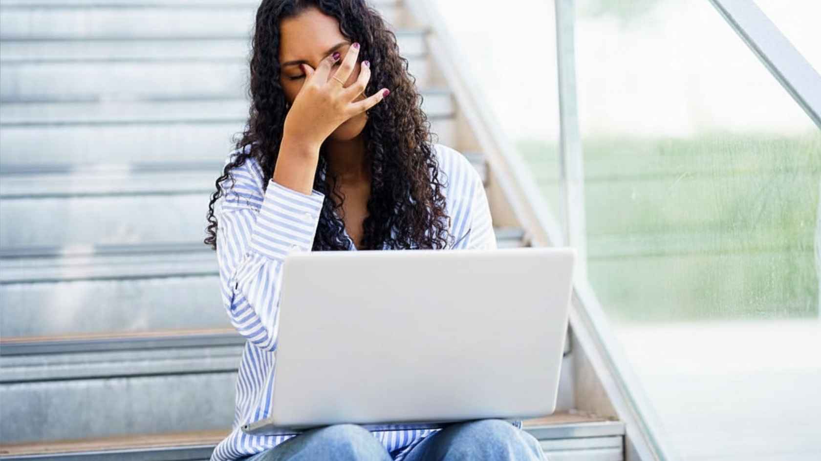 Stressed young woman rubbing her face while working on a laptop on a staircase