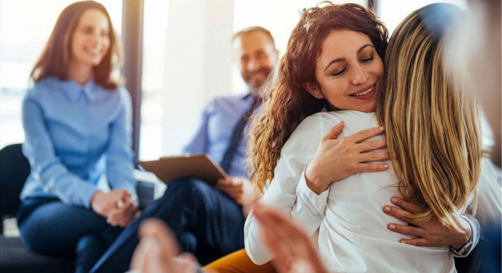 Two women embracing during a support group as others smile and applaud