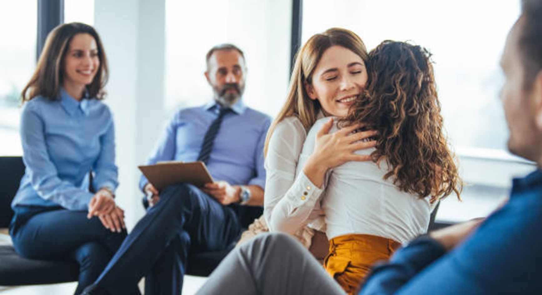 Two women embracing warmly during a group therapy session while others look on