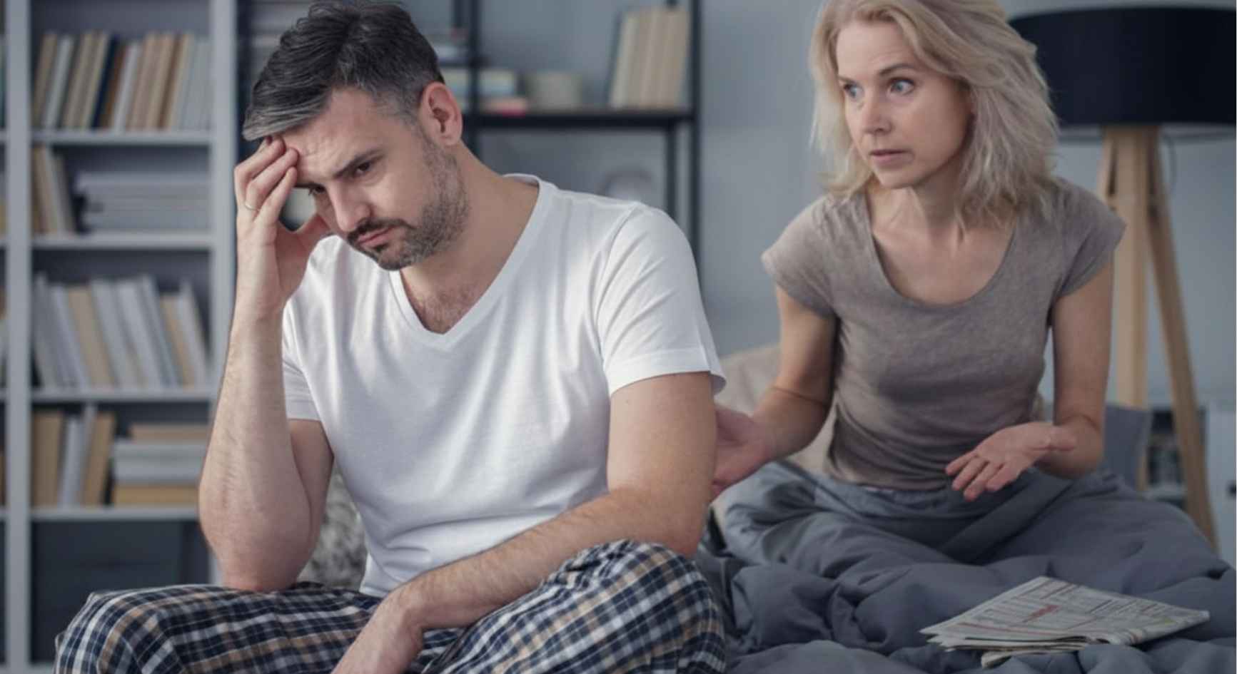 Upset man sitting on the bed while his wife argues with him in the background