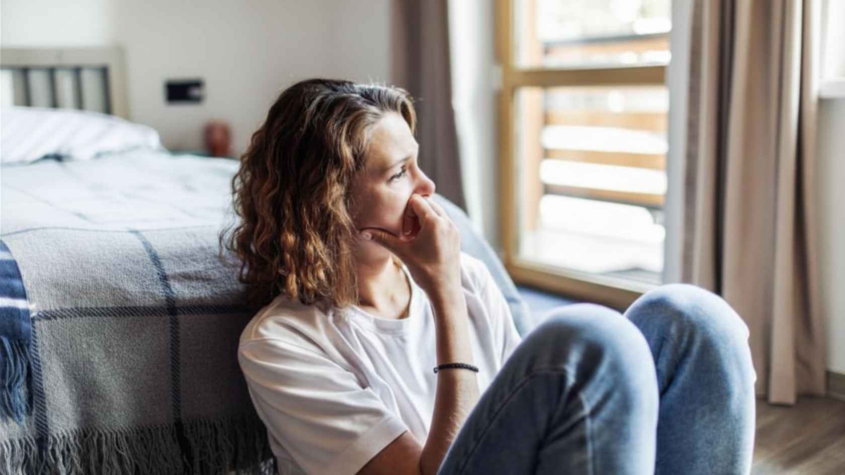 Woman sitting on bedroom floor looking out window appearing withdrawn