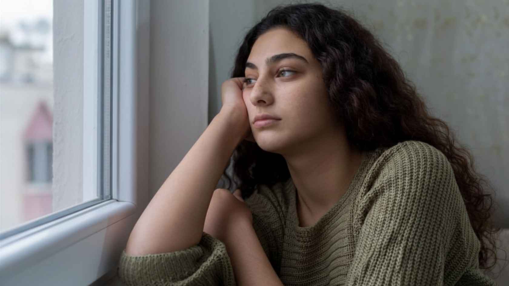 Young woman resting chin on hand gazing out window looking pensive