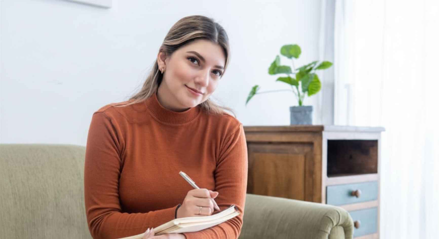Young woman writing in a notebook while sitting on a couch at home